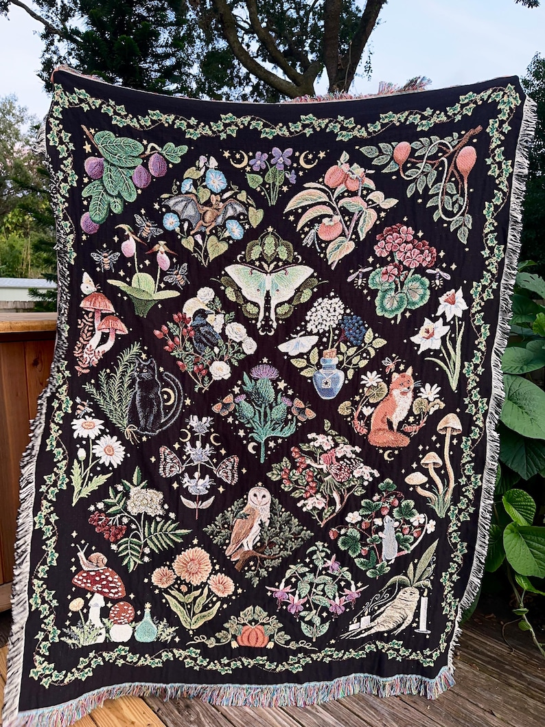 May include: A black woven throw blanket with a tapestry design featuring a variety of woodland creatures and plants, including owls, foxes, mushrooms, butterflies, and flowers. The blanket has a fringed edge.