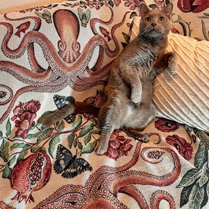 May include: A woven throw blanket featuring a detailed pattern of large orange octopuses, red pomegranates, green leaves, and black butterflies on a cream background, with a solid cream-colored textured pillow resting on top of the fabric.