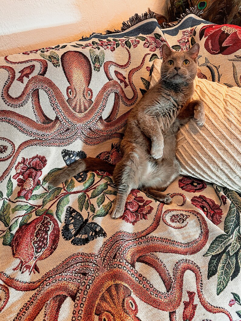 May include: A woven throw blanket featuring a detailed pattern of large orange octopuses, red pomegranates, green leaves, and black butterflies on a cream background, with a solid cream-colored textured pillow resting on top of the fabric.