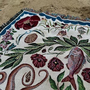 May include: A woven throw blanket featuring a cream-colored background with a detailed botanical and marine life pattern. The design includes dark red flowers, green foliage, an octopus tentacle, a fish, a seashell, and a blue border with fringe.