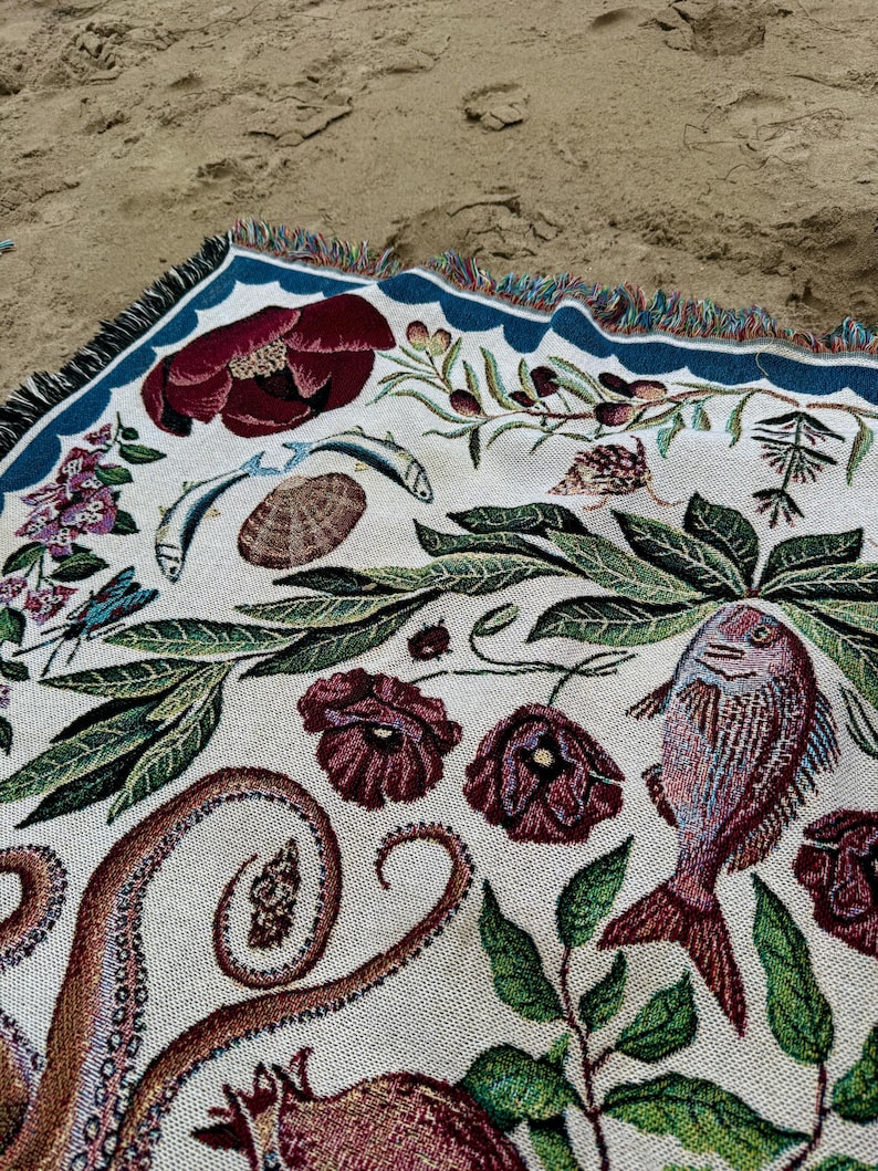 May include: A woven throw blanket featuring a cream-colored background with a detailed botanical and marine life pattern. The design includes dark red flowers, green foliage, an octopus tentacle, a fish, a seashell, and a blue border with fringe.