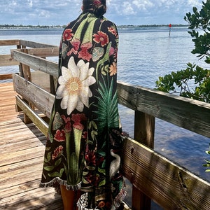May include: A woman stands on a wooden dock wearing a black and green tapestry throw blanket with a floral design. The blanket features a large white flower, green leaves, and red and pink flowers. The woman is looking out at the water.