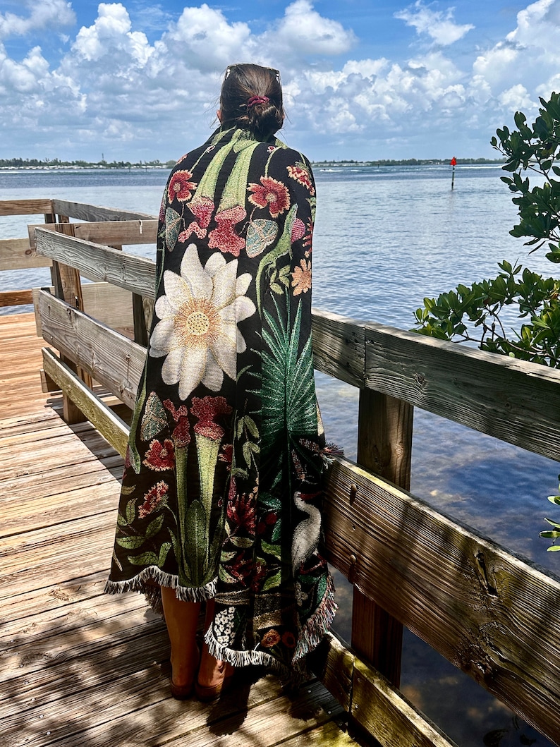 May include: A woman stands on a wooden dock wearing a black and green tapestry throw blanket with a floral design. The blanket features a large white flower, green leaves, and red and pink flowers. The woman is looking out at the water.