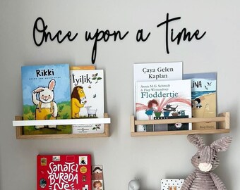 May include: A set of three wooden shelves with white metal brackets, each shelf holding a variety of children's books. The shelves are mounted on a light gray wall with the text "Once upon a time" written in black above the shelves. A gray stuffed bunny sits on the middle shelf.
