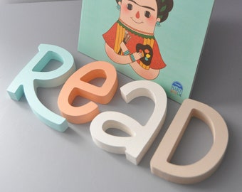 Puede incluir: Un libro infantil azul turquesa titulado "Frida Kahlo: Pequeños humanos y grandes sueños" con una ilustración de Frida Kahlo sosteniendo pinceles y una paleta. El libro está apoyado en letras de madera que deletrean "LEER".