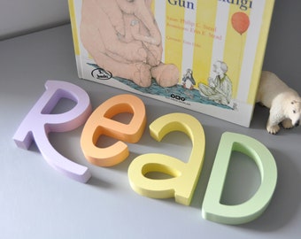 Puede incluir: Un libro infantil titulado "Amos'un Hastalandığı Gün" con una ilustración de un elefante. La palabra "READ" está escrita con letras de madera de colores pastel. Una pequeña figura de oso polar asoma por detrás del libro.