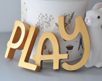 Könnte beinhalten: Goldene Holzbuchstaben, die "PLAY" buchstabieren, mit einer weißen Hasenfigur daneben.