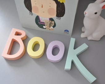 Puede incluir: Letras de madera de color pastel que deletrean la palabra "BOOK" sobre una superficie gris. Una figurita de conejo blanco está en el fondo.
