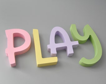Könnte beinhalten: Das Wort "PLAY" in bunten, hölzernen Buchstaben. Die Buchstaben sind rosa, gelb, lila und grün.