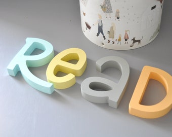 Puede incluir: Un conjunto de cuatro letras de madera que deletrean la palabra "READ" en colores pastel. Las letras son azul claro, amarillo, gris y naranja.
