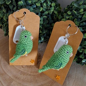 Op de afbeelding: Twee groene gehaakte papegaai sleutelhangers met gele en bruine accenten worden getoond op bruine labels. De sleutelhangers hebben zilveren ringen en witte labels. De achtergrond heeft groen gebladerte en een houten oppervlak.