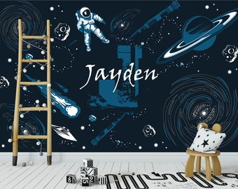 Könnte beinhalten: Eine dunkelblaue Wandmalerei zum Thema Weltraum mit einem Astronauten, Planeten und dem Namen "Jayden" in weißer Schrift. Eine Holzleiter mit Lichterketten lehnt an der Wand. Ein kleiner Holzstuhl mit einem kissen mit Sternenmuster steht auf einem Teppich.