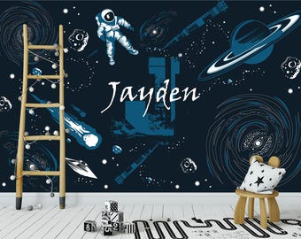 Könnte beinhalten: Eine dunkelblaue Wandtapete mit Weltraummotiv, die einen Astronauten, einen Planeten mit Ringen, Galaxien und Sterne zeigt. Der Name Jayden ist mittig in Weiß aufgedruckt. Eine Holzleiter mit Lichterkette und ein kleiner Holzstuhl stehen davor.