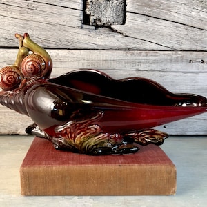 May include: A burgundy ceramic seashell planter with two green and brown snails on top. The planter is sitting on a burgundy book.