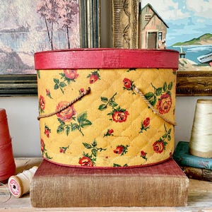 May include: A vintage yellow sewing box with a red rim and rose pattern. The box has a quilted design and rope handles. Pink and white spools of thread are on a wooden surface, with framed paintings in the background.