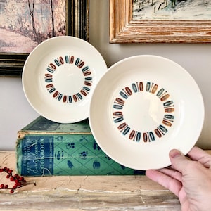 May include: Two white ceramic bowls with a blue, orange, and brown geometric pattern around the rim. The bowls are stacked on top of a green cookbook.