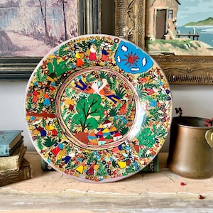 May include: A decorative ceramic plate with a vibrant folk art design. The plate features a central image of two birds, trees, and small houses, surrounded by a border of figures and floral patterns. The plate has a blue accent with a sun design.