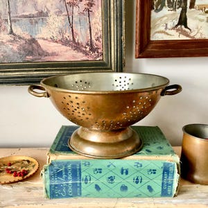 May include: A vintage copper colander with a stand and holes throughout. The colander is sitting on top of a green and white cookbook.