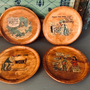 Puede incluir: Conjunto de cuatro posavasos de madera con ilustraciones humorísticas y texto. Los posavasos muestran a un hombre cortando muñecas de papel, un hombre al teléfono, un hombre pensando y una mujer sentada con un grupo de personas. El texto en los posavasos incluye "Cut out smokin", "Cut out drinkin", "Cut out winman", "Don't think woik", "Sumfah towd" y "Whatcha mean "momber please?"
