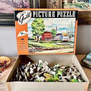 Puede incluir: Una caja de cartón abierta que contiene un rompecabezas de imágenes Guild vintage con 304 piezas. La caja del rompecabezas es naranja y presenta una colorida escena de paisaje con casas, árboles y una iglesia. La caja está etiquetada como "Guild Picture Puzzle" en texto negro.