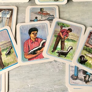 May include: A set of illustrated cards featuring a skunk in various situations. The cards are white with blue borders and depict a skunk being held by a person, a skunk walking in a yard, a skunk near a house, and a skunk near a water fountain.