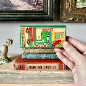 May include: A vintage toy garage with a green border, featuring illustrations of cars and a garage building. The toy is held over a stack of old books, including one titled "Bedtime Stories". A ceramic rooster figurine is also visible.