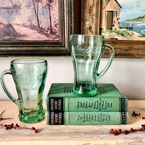 Könnte beinhalten: Zwei grüne Vintage-Coca-Cola-Tassen mit Griffen. Das Coca-Cola-Logo ist auf der Vorderseite jeder Tasse zu sehen. Die Tassen stehen auf zwei grünen Büchern. Der Hintergrund umfasst gerahmte Gemälde.
