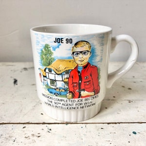 May include: White ceramic mug featuring a colourful illustration of a boy wearing glasses and a red jacket. The text on the mug reads "Joe 90 Mission Completed Joe Becomes The 90th Agent for WIN World Intelligence Network".