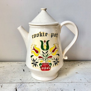 Puede incluir: Jarrón de galletas de cerámica blanca con forma de tetera con un diseño floral y las palabras "cookie pot" en la parte delantera.