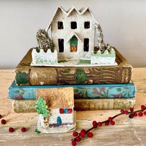Puede incluir: Una exhibición de estilo vintage con una casa de papel blanca con un techo texturizado, colocada sobre una pila de libros antiguos. Los libros son de varios colores, con cubiertas decorativas. Una pequeña casa de papel y bayas rojas completan la escena.