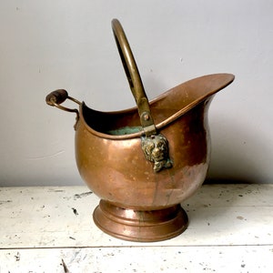 May include: A vintage copper coal scuttle with a brass handle and lion head finial. The scuttle has a rounded body and a wide base.