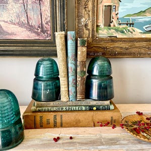 Op de afbeelding: Een opstelling van vintage boeken, teal-groene glasisolatoren en een decoratief houten bord met rode bessen. De boeken hebben verouderde covers. Twee ingelijste schilderijen zijn op de achtergrond.