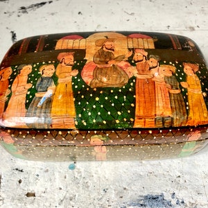 Może przedstawiać: Prostokątne drewniane pudełko z ręcznie malowanym obrazem przedstawiającym osoby w tradycyjnych strojach. Pudełko jest ozdobione zielonym i złotym geometrycznym wzorem.