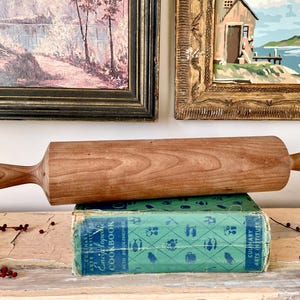 May include: A wooden rolling pin with rounded handles rests on a vintage cookbook. The rolling pin is a light brown color and has a smooth finish. The cookbook is green and blue with text.