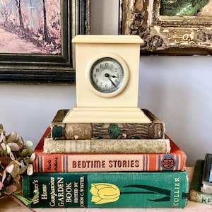 May include: A cream-colored vintage clock with a round face sits atop a stack of antique books. The clock has a square base and a small, circular clock face. The books are in various colors, including green, orange, and brown. The words "BEDTIME STORIES" are visible on one book.