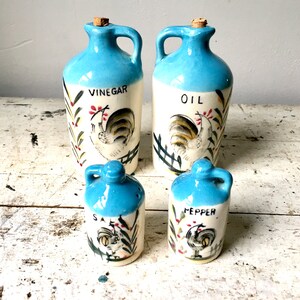 Op de afbeelding: Set van vier blauwe keramische zout- en peperstrooiers en olie- en azijnflessen. De flessen hebben een witte achtergrond met een haanontwerp en de woorden "olie", "azijn", "zout" en "peper" erop.