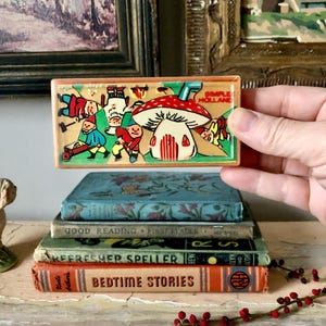 Può includere: Un giocattolo rettangolare vintage in legno con un'illustrazione colorata di gnomi e una casa a forma di fungo. Il giocattolo è tenuto sopra una pila di libri antichi, tra cui "Bedtime Stories" e "Good Reading". Il testo "SIMPLE HOLLAND" è visibile.