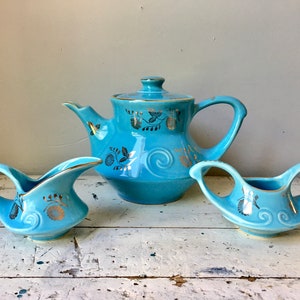 May include: A blue teapot and two creamer pitchers with gold floral designs. The teapot has a rounded body and a spout on the side. The creamers have a curved handle and a spout on the side.