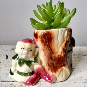 Op de afbeelding: Een keramische plantenpot in de vorm van een boomstronk met een klein elfje ernaast. De pot is versierd met bruine en witte glazuur en heeft een groene succulent plant erin.