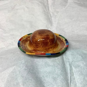 Puede incluir: Un recipiente de mantequilla de cerámica marrón con un borde colorido. El recipiente tiene una parte superior redondeada y una base ancha. El borde está decorado con rayas rojas, verdes, azules y amarillas.
