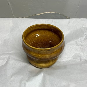 Puede incluir: Un pequeño cuenco de cerámica marrón con un esmalte moteado. El cuenco tiene un fondo redondeado y un borde ligeramente acampanado.