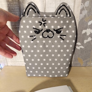 Peut inclure: Une pochette en tissu gris avec un motif d'étoiles blanches. La pochette est en forme de chat avec des yeux, un nez et des moustaches brodés en noir. Le chat a une expression fâchée avec un froncement de sourcils brodé en noir.