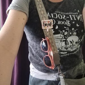 Peut inclure: Une bandoulière de sac à motifs beiges avec un clip rectangulaire blanc orné d'un chat de dessin animé portant des lunettes de soleil noires. Une paire de lunettes de soleil à monture rose est suspendue au clip. Le sac gris est porté sur un t-shirt.