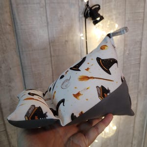 Peut inclure: Un support de livre sur le thème d'Halloween, blanc et gris, avec un motif de chapeau de sorcière, de balai et de bougie. Le support a un fond gris et une boucle pour le transport. Le petit support a le même motif.