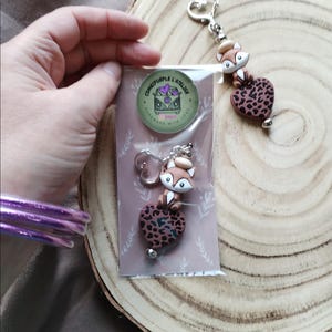 Peut inclure: Un porte-clés et une paire de boucles d'oreilles avec un motif de renard. Le renard est perché sur un charm en forme de cœur avec un motif léopard. Le porte-clés a un fermoir en forme de cœur. Les boucles d'oreilles sont emballées dans un sac en plastique transparent.