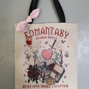 Peut inclure: Un sac fourre-tout en toile couleur crème avec des anses noires et une breloque nœud rose pâle. L'avant présente un motif avec le texte ROMANTASY READERS SOCIETY et JUST ONE MORE CHAPTER, illustrant des livres, un cœur, une épée et des roses.