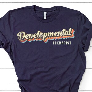 May include: A navy blue t-shirt with a retro-style graphic that says "Developmental Therapist" in yellow text with a rainbow-colored background.