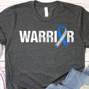K&ouml;nnte beinhalten: Ein dunkelgraues T-Shirt mit dem Wort "WARRIOR" in wei&szlig; und einem Used-Look-Effekt bedruckt. Auf der rechten Seite des Shirts ist ein blaues Band mit dem Text "TYPE 1 DIABETES" aufgedruckt.