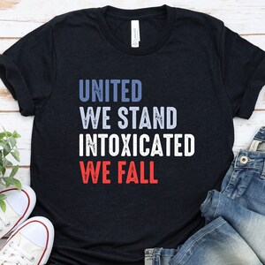 May include: A black t-shirt with the text "UNITED WE STAND INTOXICATED WE FALL" in blue, white, and red. The shirt is paired with blue jeans and white sneakers.
