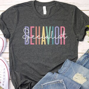 May include: A dark heather grey t-shirt with the text "BEHAVIOR Specialist" in rainbow colors. The shirt is folded and laid on a white wooden surface with a pair of white sneakers and blue jeans.